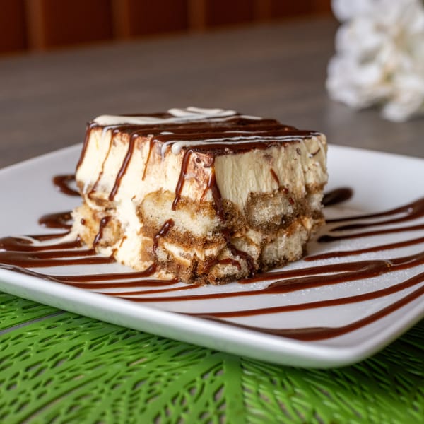 A slice of tiramisu with chocolate and white sauce drizzle on a white plate with a green placemat.
