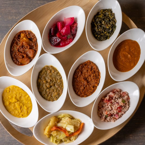 Nine small white oval dishes filled with various colorful Ethiopian stews and vegetable sides on a wooden tray.