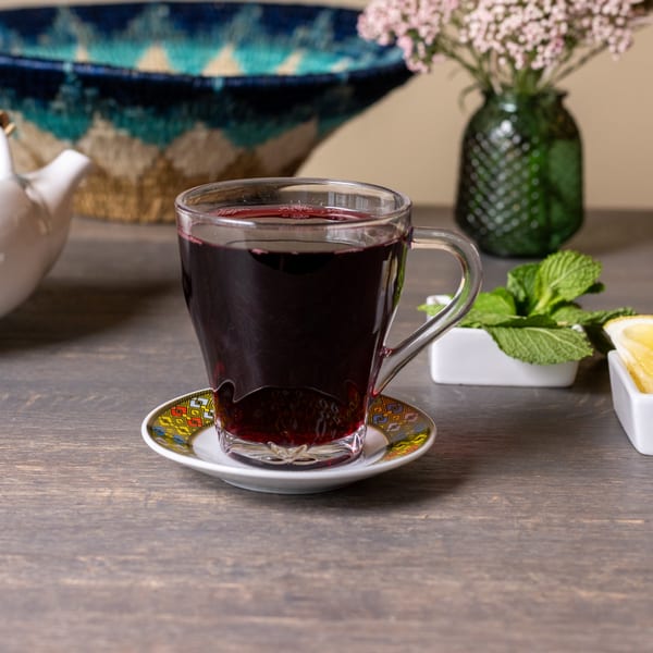 A glass of dark red Ethiopian tea on a colorful saucer, with mint and lemon nearby.