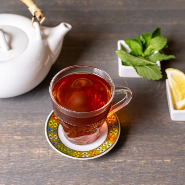 A glass of Ethiopian tea on a colorful saucer, with a white teapot, mint, and lemon.