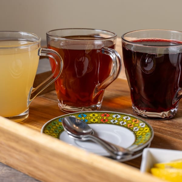 Three clear mugs with different colored drinks, a spoon on a patterned saucer, and a wooden tray.