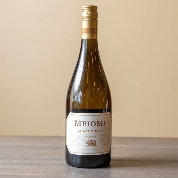 A bottle of Meiomi Chardonnay wine with a gold label and cap stands on a wooden surface.