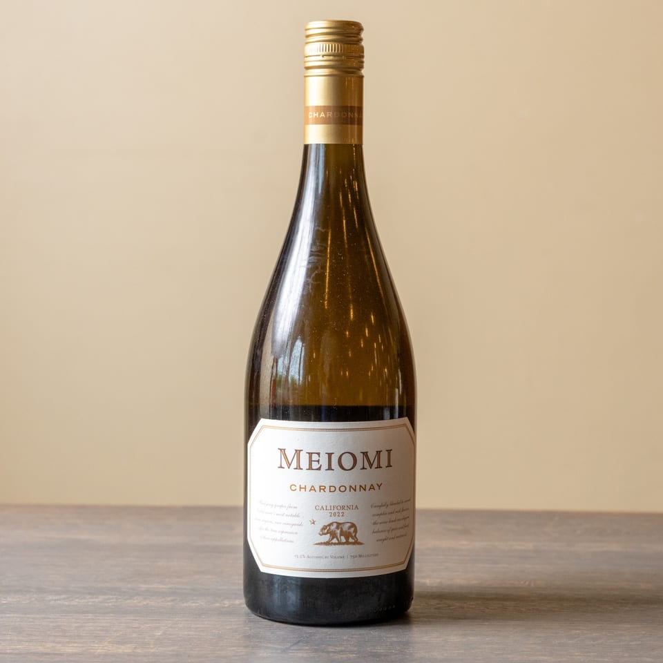 A bottle of Meiomi Chardonnay wine with a gold label and cap stands on a wooden surface.