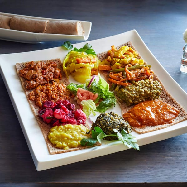 A white platter of colorful Ethiopian dishes, including injera, stews, and vegetables, with rolled injera in the background.