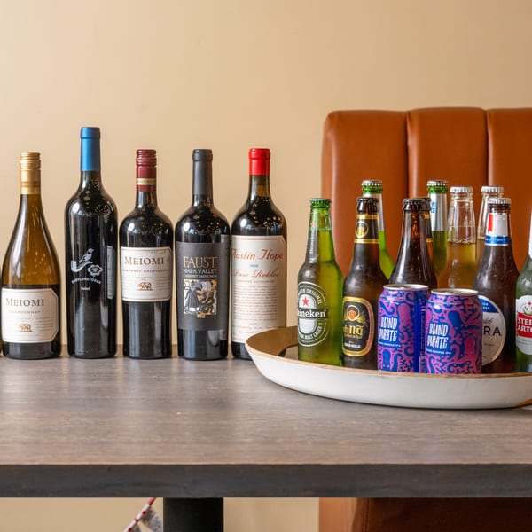 A selection of wine bottles, beer bottles, and beer cans on a wooden table with a brown booth in the background.
