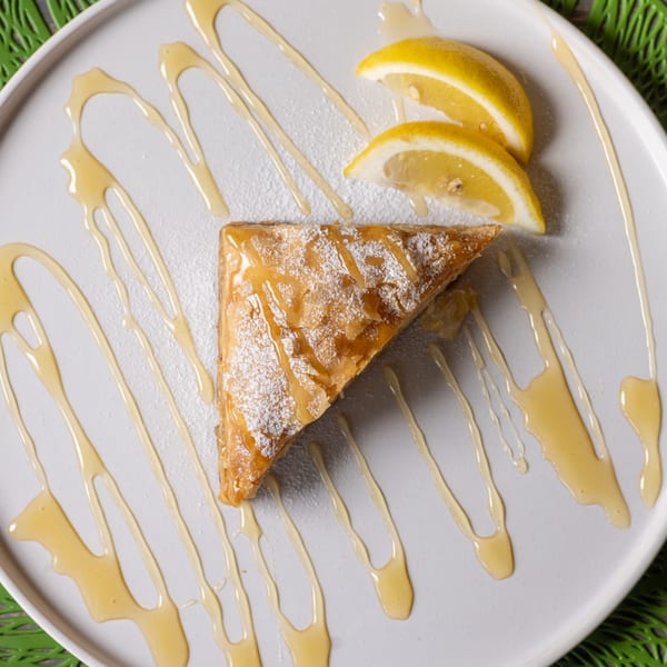 A triangular pastry drizzled with honey and powdered sugar, served with two lemon wedges.
