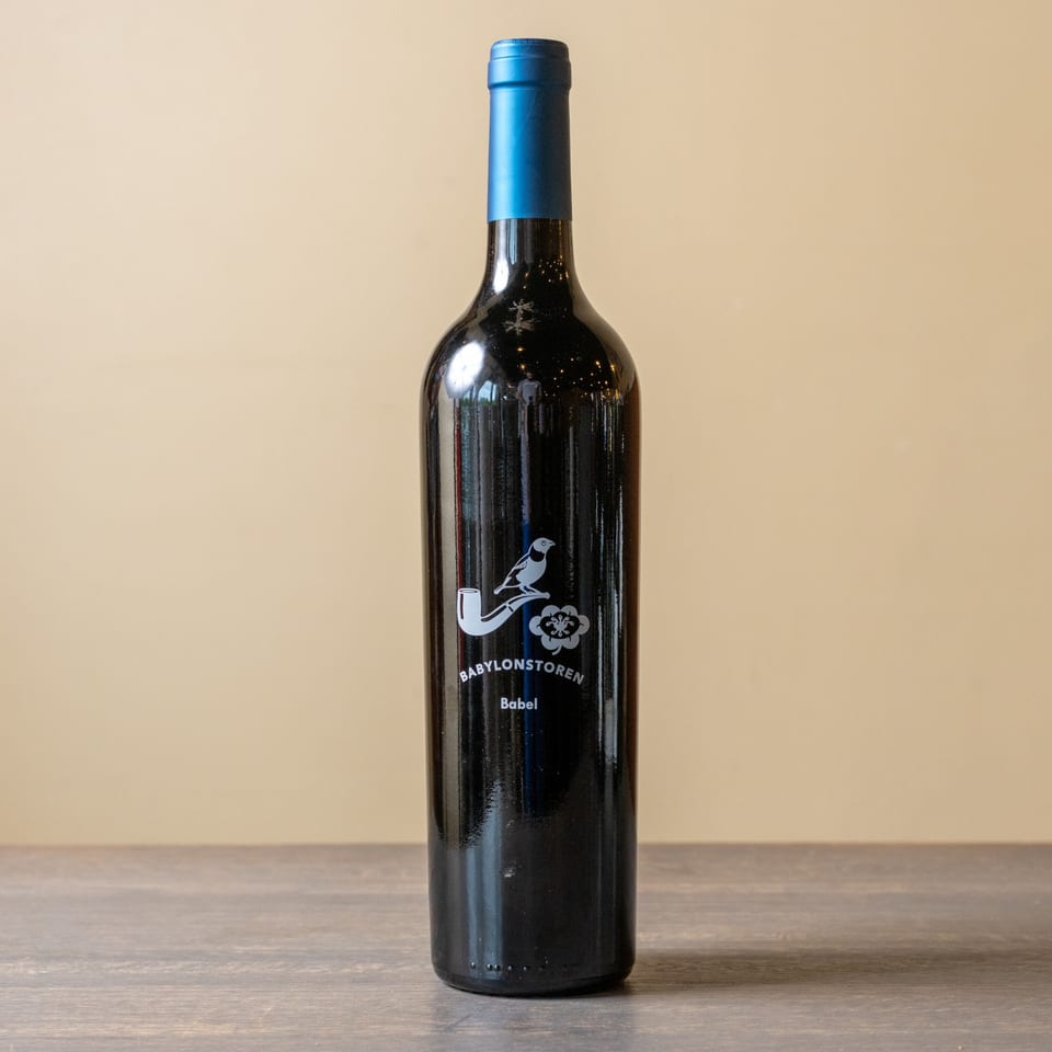 A dark wine bottle with a blue foil top and a white bird-on-pipe logo stands on a wooden table.