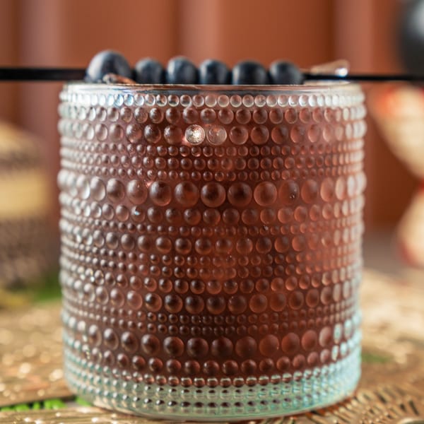 Close-up of a textured glass filled with a dark beverage, garnished with blueberries and a black straw.