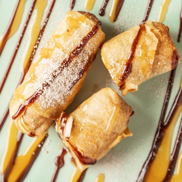 Three golden-brown spring rolls, drizzled with caramel and chocolate sauce, dusted with powdered sugar.